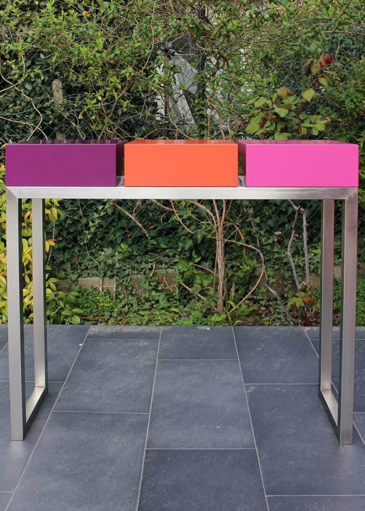 Console moderne décorative avec 3 tiroirs sur mesure à ouverture en façade. Console Pied-Estal avec un piétement en inox brossé. Tiroirs en couleurs finition laque contemporaine satinée, couleurs aubergine, orange pop, fuchsia. Photo vue d'ensemble