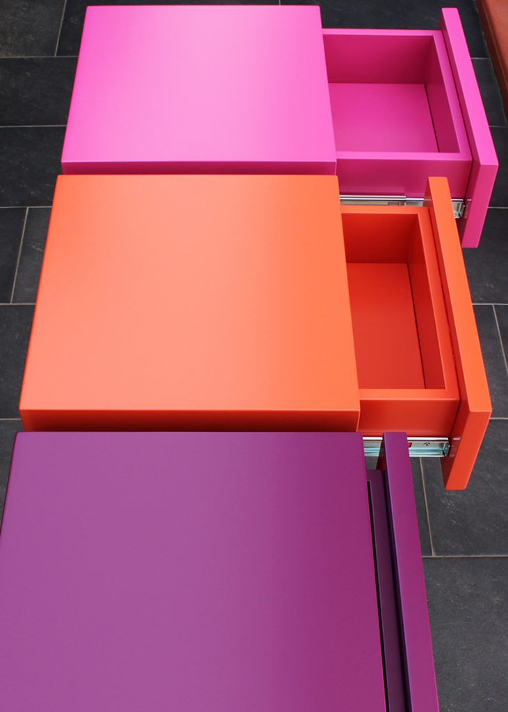 Console moderne décorative avec 3 tiroirs sur mesure à ouverture en façade. Console Pied-Estal avec un piétement en inox brossé. Tiroirs en couleurs finition laque contemporaine satinée, couleurs aubergine, orange pop, fuchsia. Photo détail tiroirs ouverts