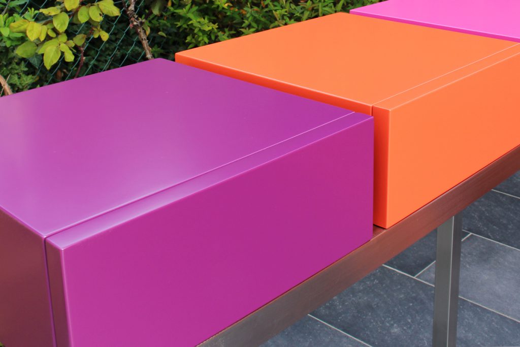 Console moderne décorative avec 3 tiroirs sur mesure à ouverture en façade. Console Pied-Estal avec un piétement en inox brossé. Tiroirs en couleurs finition laque contemporaine satinée, couleurs aubergine, orange pop, fuchsia. Photo détail vue avant